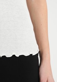 Camiseta de tirantes blanca con costillas y dobladillo festoneado, con una textura sutil, combinada con tejido negro en la parte inferior. Sin herrajes visibles ni patrones.