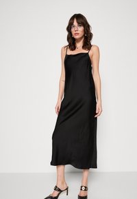 Robe de satin noir à bretelles fines, texture lisse et design fluide qui atteint le milieu du mollet. Associée à des mules noires à talons.