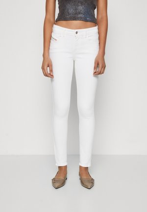 Femme portant un jean skinny blanc et un crop top gris foncé sans manches, debout avec les bras détendus le long du corps sur un fond uni.