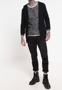 Sudadera con cremallera negra sobre una camisa de manga larga gris, combinada con jeans rasgados negros y botas negras de cordones. Presenta tatuajes en los brazos.