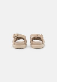 Beige sandaalit, joissa on sileä pinta, avoin takaosa ja säädettävä solkiremmi. Pehmeä pohja kevyellä, teksturoidulla ulkopohjalla.