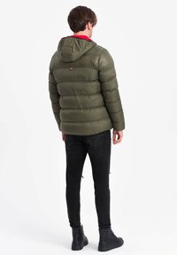 Grön puffjacka med huva, quiltad textur, röd innerfoder, bärs med svarta jeans och svarta stövlar, sedd bakifrån.