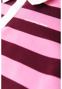 Gros plan d'une chemise rayée rose et rouge foncé avec un col blanc et le texte « COLOURFUL REBEL » brodé sur la poitrine.