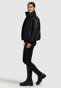 Kvinde, der står i en sort polstret jakke, sorte leggings og knæhøje sorte støvler mod en ensfarvet lys baggrund.