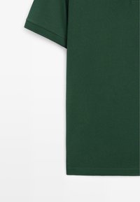 Chemise vert foncé à manches courtes sur fond blanc, montrant le côté supérieur gauche et les coutures de l'ourlet.