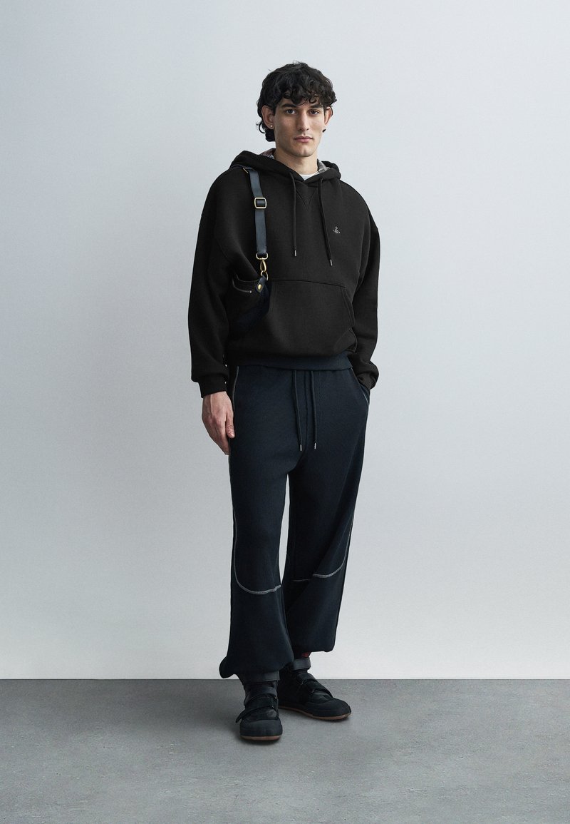 Sweat à capuche noir avec cordons, associé à un pantalon ample bleu foncé côtelé avec des coutures claires. Porté avec des baskets noires et un sac à bandoulière.