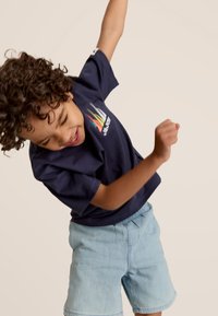 Marineblauwe katoenen t-shirt met kleurrijk grafisch ontwerp en korte mouwen, gecombineerd met lichtblauwe denimshorts. Kind in speelse houding.