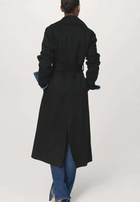 Manteau en laine noir avec une taille ceinturée, des manches longues et une fente au dos. Associé à un jean en denim bleu avec des ourlets effilochés.