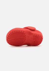 Punainen kumisandaali, jossa on kuvioitu pohja, säädettävä remmi ja sileä yläosa. Varustettu kohotetulla logolla ja kuvioidulla kulutuspinnalla pitoa varten.