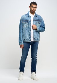 Chaqueta de denim azul claro con dos bolsillos en el pecho, cierre de botones y textura desgastada, combinada con jeans oscuros ajustados y zapatillas blancas.