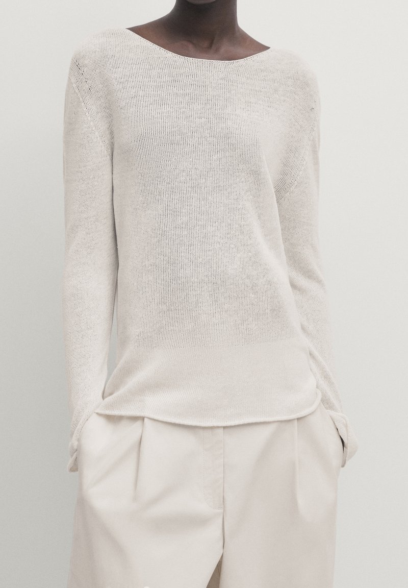 Pull beige clair en maille avec un large col bateau et des manches longues, présentant une coupe ample et une surface texturée. Associé à un pantalon crème.