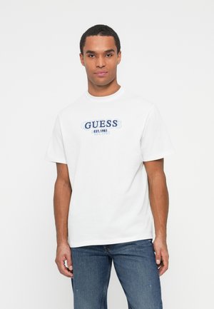 Hombre vestido con una camiseta blanca de manga corta que dice "GUESS EST. 1981" en texto azul, combinada con jeans azules, frente a un fondo blanco liso.