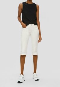 Valkoiset caprit, jotka on kääritty ylös, yhdistetty mustaan kuitupintaiseen hihattomaan toppiin. Malli käyttää valkoisia tennareita, mikä korostaa rentoa tyyliä.