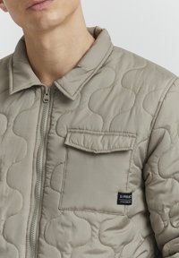 Beige quiltet jakke med glidelås foran, spiss krage og en brystlomme med merkeetikett. Myk, strukturert stoff med en glatt overflate.