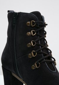 Czarne zamszowe botki za kostkę z bocznym zamkiem, sznurowane z przodu z mosiężnymi oczkami oraz wysokim słupkowym obcasem. Gładka struktura i przemyślany design.