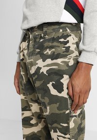 Calças cargo de camuflagem com um padrão mesclado de verde, castanho e bege, com um bolso lateral e tecido resistente.
