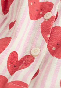 Pamutanyagból készült ing, amely nagy piros szíveket ábrázol rózsaszín és fehér csíkos háttéren, puha tapintású, kis rózsaszín gombbal.
