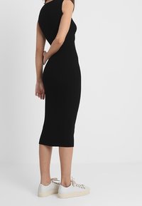 Femme portant une robe midi noire ajustée et sans manches, ainsi que des baskets blanches, debout avec les mains derrière le dos sur un fond uni.