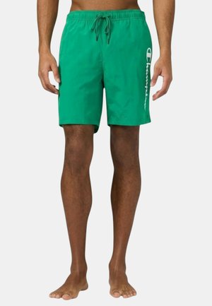 Uomo che indossa pantaloncini da bagno verdi con logo bianco Champion sulla gamba sinistra, in piedi a piedi nudi su uno sfondo semplice.