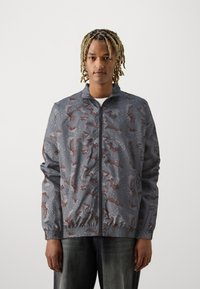 Vandafvisende grå jakke med et brunt camouflage-mønster, der har lynlås foran, elastiske manchetter og en opretstående krave. Glat tekstur.