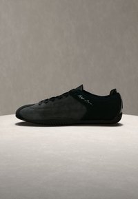 Μαύρο sneaker χαμηλού προφίλ με επάνω μέρος από σουέντ, δέσιμο με κορδόνια μπροστά, χαρακτηριστική λεπτομέρεια στο πλάι και λεπτή καουτσούκ σόλα σε ουδέτερη επιφάνεια.
