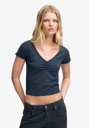 Mujer rubia con una camiseta ajustada azul marino de manga corta y escote en V y pantalones oscuros de pana, de pie con las manos en los bolsillos.