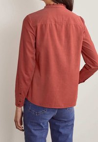 Camicia a maniche lunghe di colore rosso chiaro con colletto arricciato, polsini abbottonati e texture liscia. Abbinata a jeans blu. Vista posteriore mostrata.