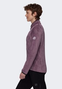 Chaqueta de forro polar de color morado claro con un patrón texturizado, cuello alto y detalles en negro; presenta un logotipo en la manga.