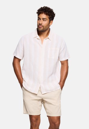 Hombre con camisa beige clara de manga corta abotonada y pantalones cortos beige a juego, con las manos en los bolsillos, mirando hacia un lado.