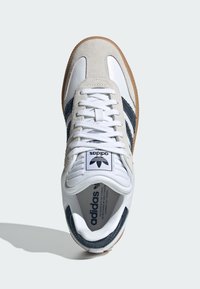 Fehér sneaker tengerészkék csíkokkal, velúr és bőr anyagból, kerek orral, fűzős elejével és gumírozott talppal. A márkázott nyelv címkéje is jól látható.