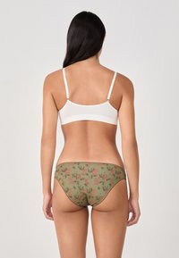 Woman seen from the back wearing a white bra and green patterned briefs with pink floral designs, standing against a plain light background.