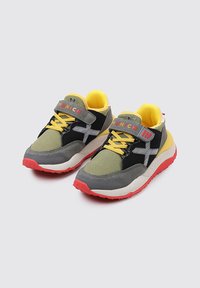 Zapatillas deportivas en gris y negro con acentos en amarillo, material de malla y gamuza, con cordones y tira de Velcro, suela de goma roja con un patrón.