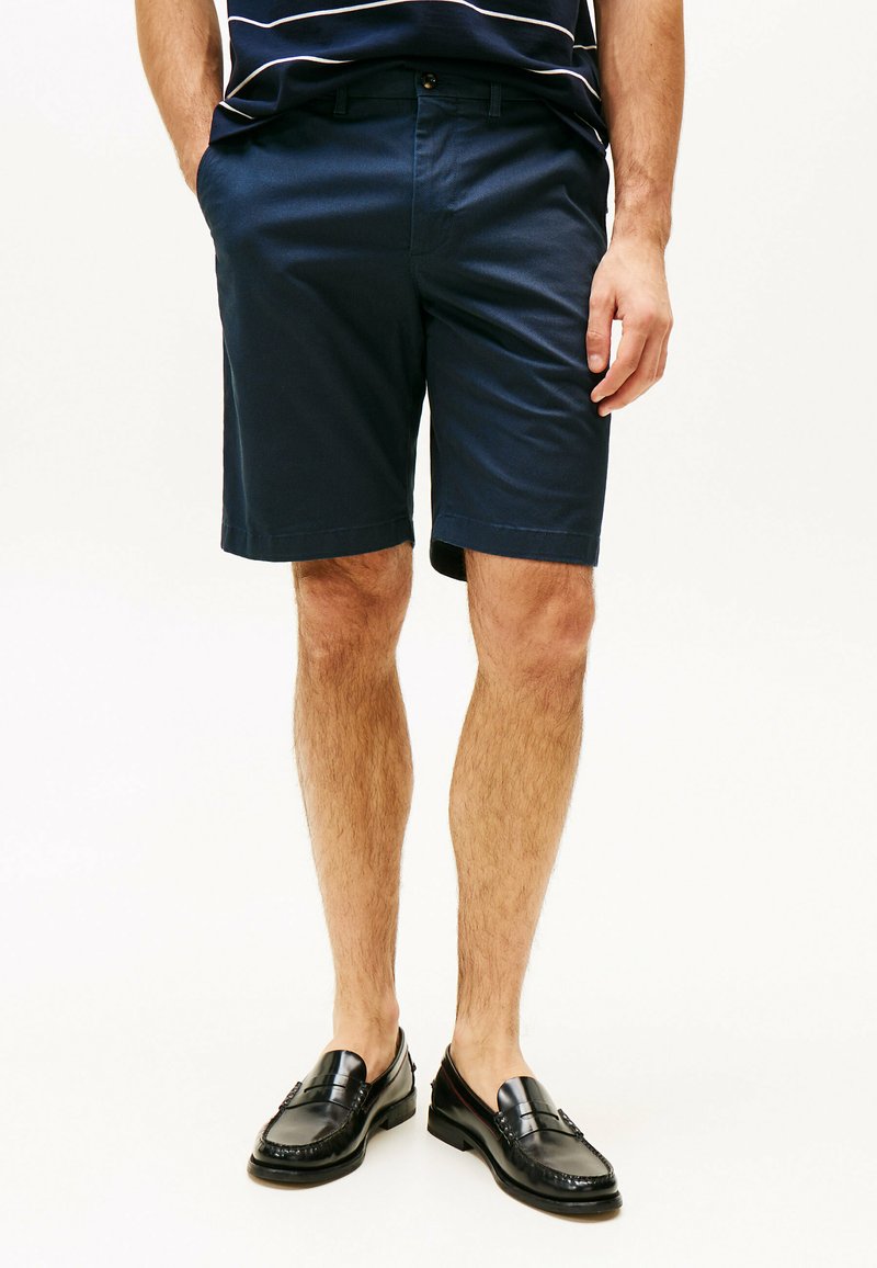 Pantaloni corti in cotone blu navy con due tasche, taglio dritto e chiusura con bottone. Indossati con mocassini neri su gambe scoperte.