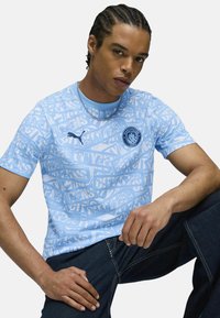 Ljust blå kortärmad t-shirt med ett grafiskt tryck med texten "CITYZENS" och en cirkulär Manchester City-logotyp. Tillverkad av en poly-bomullsblandning.