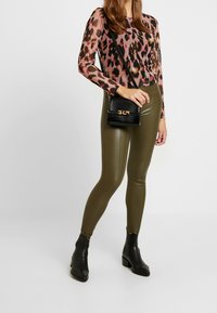 Langärmliges Top mit Leopardenmuster in Pink und Schwarz, kombiniert mit olivgrünen Leggings und schwarzen Ankle Boots. Accessoire: kleine schwarze Umhängetasche.