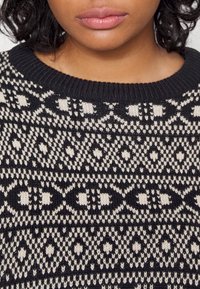 Gros plan sur le bas du visage et le cou d'une personne portant un pull en tricot noir et blanc à motifs géométriques.