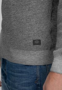 Grauer Strickpullover mit strukturiertem Muster, gerippten Bündchen und Saum. Verfügt über ein kleines schwarzes Etikett auf der unteren linken Seite. Wird mit blauen Jeans kombiniert.