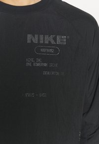 Svart langermet skjorte med teksturert stoff. Har sølvtrykt merkevare og tekst, inkludert Nike-logoen og adresseopplysninger.