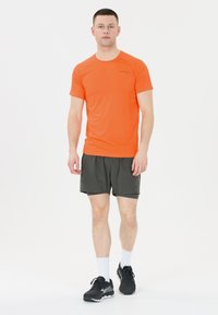 Kirkas oranssi urheilupaita lyhyillä hihoilla, yhdistettynä tummanvihreisiin shortsisiin. Mallilla on päällään mustat juoksukengät ja valkoiset sukat.