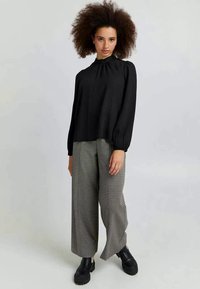 Schwarze Bluse mit langen Ärmeln und gerafftem Halsdetail, kombiniert mit weit geschnittenen Hosen im Hahnentrittmuster und klobigen schwarzen Stiefeletten.