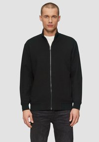 Schwarze Zip-Jacke aus strukturiertem Stoff, hoher Kragen und gerippte Bündchen. Kombiniert mit dunklen Jeans, was einen minimalistischen Look kreiert.
