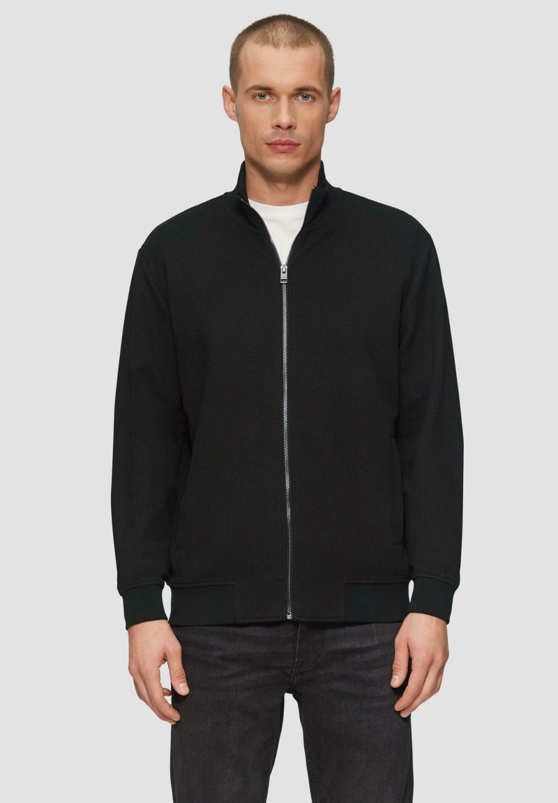 Schwarze Zip-Jacke aus strukturiertem Stoff, hoher Kragen und gerippte Bündchen. Kombiniert mit dunklen Jeans, was einen minimalistischen Look kreiert.