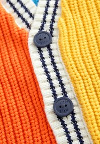 Bunt gestrickte Strickjacke mit orangefarbenen, gelben und cremefarbenen Streifen, mit einem marineblauen Knopf, der "Little Bird" beschriftet ist. Strukturiertes Gewebedetail.