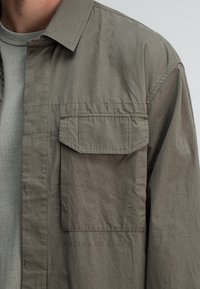 Man in olijfgroene overhemd met knoopsluiting en een flapzak op de borst, met vervaagde tekst "BUTCHER OF BLUE J" onder de zak.