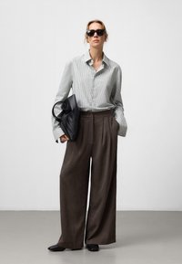 Camicia a righe nei toni del blu pallido e bianco, abbinata a pantaloni larghi marroni, con una borsa in pelle nera e occhiali da sole neri.