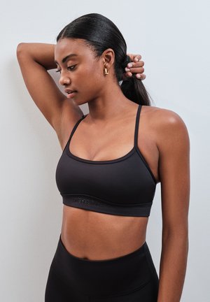 Femme aux cheveux foncés portant un soutien-gorge de sport noir et un legging, levant un bras et tenant ses cheveux en queue de cheval.