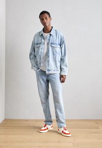 Lys blå denimjakke med to brystlommer, matchende lys blå jeans og hvite sneakers med røde detaljer og snøring.