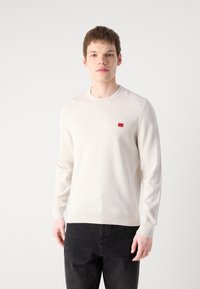 Un pull en tricot crème clair avec un col rond et un petit patch logo rouge sur la poitrine. Porté avec un jean noir, mettant en avant des lignes simples et épurées.