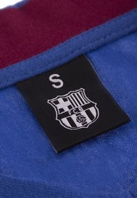 Blauwe stof met een kastanjebruine kraag. Zwarte label met een witte schild met gestreept ontwerp en "FCB" tekst, wat maat klein (S) aangeeft.