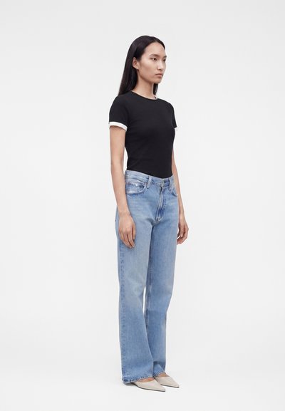 Γυναίκα στέκεται στραμμένη αριστερά, φορώντας μαύρο μπλουζάκι με λευκή λεπτομέρεια, ανοιχτό μπλε τζιν παντελόνι με ίσια πόδια και μπεζ μυτερά παπούτσια με φόντο λευκό.